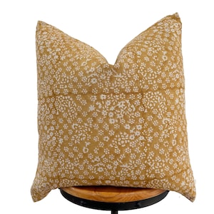 May include: A square decorative pillow with a mustard yellow background and a repeating pattern of small white floral designs. The pillow is resting on a wooden stool with a black metal base. The pillow is a home decor item.