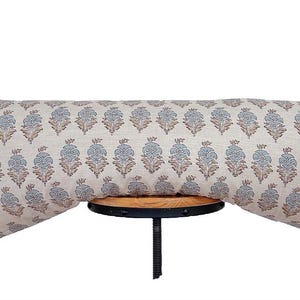 Könnte beinhalten: Ein langes, rechteckiges Zierkissen mit einem sich wiederholenden Blumenmuster in Blau und Braun auf cremefarbenem Hintergrund. Das Kissen liegt auf einem kleinen, runden Holzschemel mit schwarzem Metallsockel.