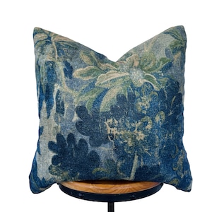 May include: A square decorative pillow with a floral pattern in shades of blue, green, and beige. The pillow has a textured appearance and is likely made of fabric. The design features large, stylized flowers.