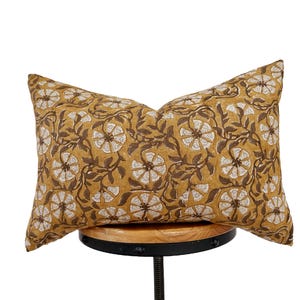 May include: Mustard-colored rectangular pillow with a floral pattern in brown and white. The pillow is placed on a wooden stool. The floral design features stylized flowers and leaves, creating a decorative home accent.