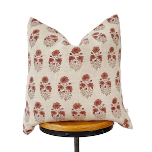May include: A beige pillow with a floral pattern in shades of red and green. The pillow is sitting on a wooden stool.