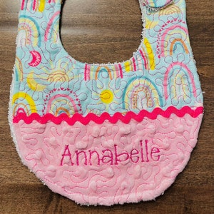 May include: A light blue baby bib with a rainbow and sun pattern. The bib has a pink, scalloped trim and a pink, soft, textured backing with the name "Annabelle" embroidered in pink thread.