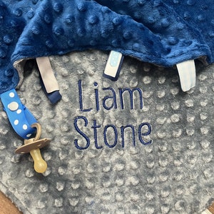 May include: A gray and blue baby blanket with a pacifier attached. The blanket has a textured surface and the name "Liam Stone" is embroidered on it.
