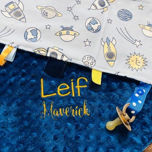 May include: A blue and white baby blanket with a space theme. The blanket has a soft, textured side and a smooth side with a print of rockets, planets, stars, and aliens. The blanket is personalized with the name "Leif Maverick" in yellow embroidery.