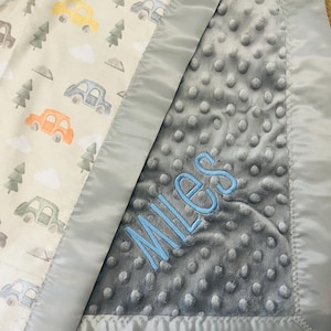 May include: A baby blanket with a car and tree pattern on one side and a gray, textured fabric on the other. The blanket has a satin border and the name "MILES" embroidered in blue.