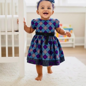 Könnte beinhalten: Ein lächelndes Baby trägt ein gemustertes Kleid in Lila, Blau und Schwarz mit passendem Stirnband und schwarzer Schleife. Das Kleid hat kurze Ärmel und einen schwarzen Gürtel. Das Baby steht auf einem weißen Teppich.