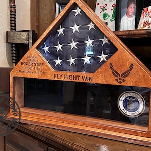May include: Wooden flag display case with a US flag, Air Force emblem, and text "FLY FIGHT WIN." Includes a space for a military coin and a nameplate with the text "SENIOR AIRMAN ROGER STIPE."