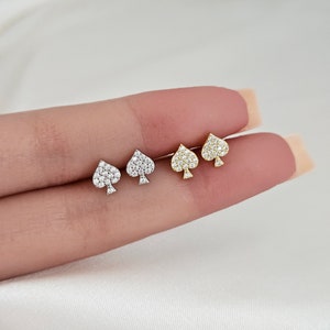 May include: Three pairs of small stud earrings in the shape of a spade, with a pave setting of clear cubic zirconia stones. The earrings are shown on a hand, with the gold pair on the left, the silver pair in the middle, and the rose gold pair on the right.