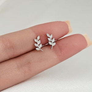 May include: A pair of silver-colored stud earrings. Each earring features a delicate leaf design with small, sparkling clear stones. The earrings are displayed on a light-colored background.