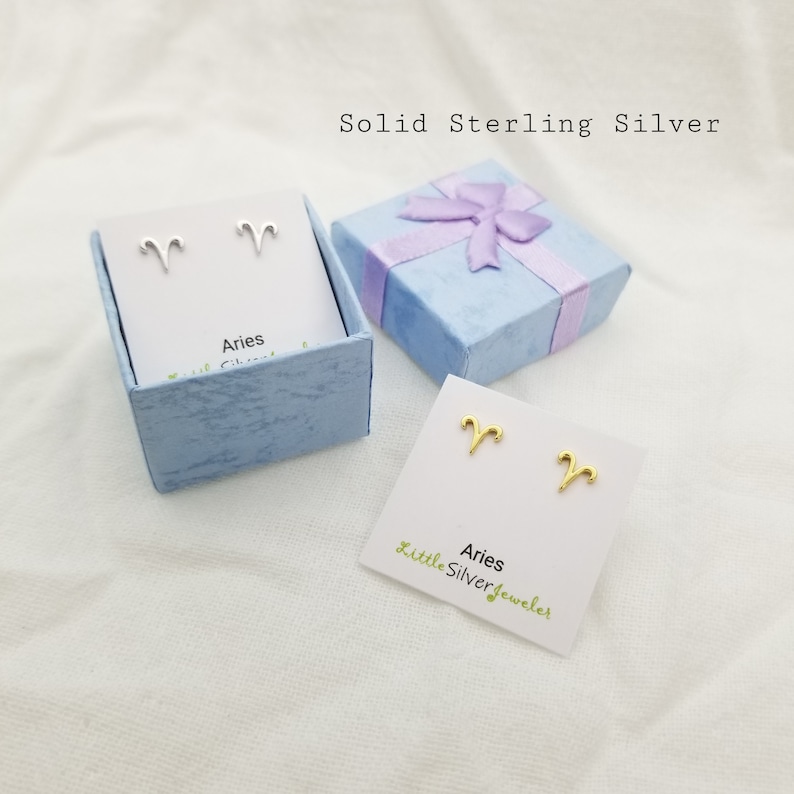 May include: A pair of silver and gold Aries zodiac sign earrings in a blue gift box. The earrings are on a white card with the text "Aries Little Silver Jeweler".