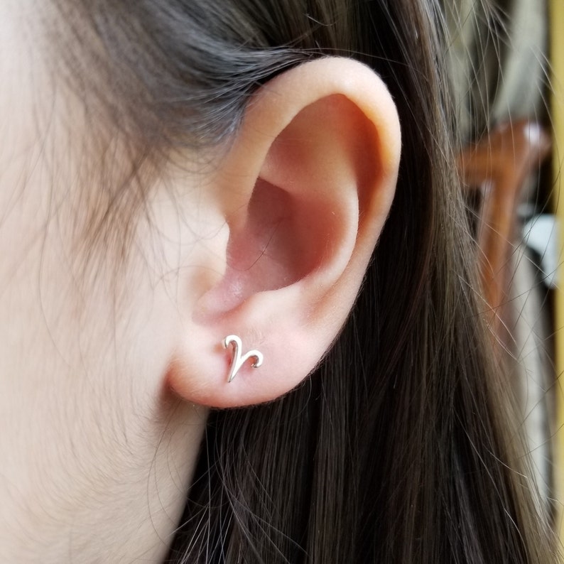 May include: A close-up of a person's ear with a silver Aries zodiac sign stud earring.