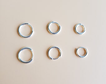 May include: Six silver hoop earrings of different sizes on a beige background.