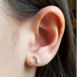 May include: A close-up of a person's ear with a silver dragonfly earring. The earring is small and delicate, with intricate details.
