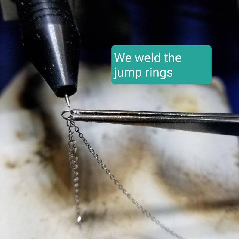May include: A close-up of a silver jump ring being welded with a torch. The jump ring is held in place with tweezers. The text "We weld the jump rings" is visible in the image.