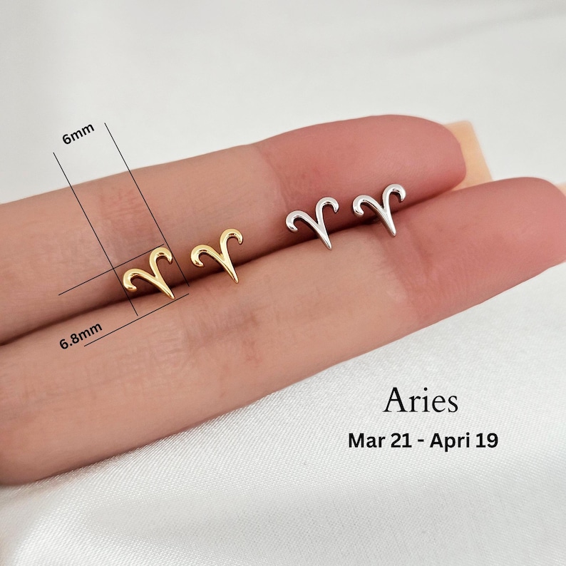 May include: Gold and silver Aries zodiac sign earrings. The earrings are 6 mm wide and 6.8 mm tall. The earrings are on a person's finger. The text "Aries Mar 21 - Apr 19" is below the earrings.