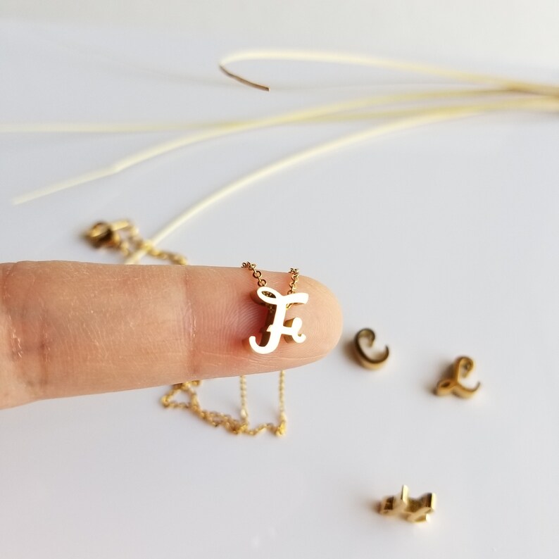 May include: A gold chain necklace with a cursive letter "F" pendant. The pendant is white and the chain is gold.