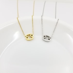 May include: Two delicate gold and silver necklaces with a Cancer zodiac sign pendant. The necklaces are displayed on a white plate.