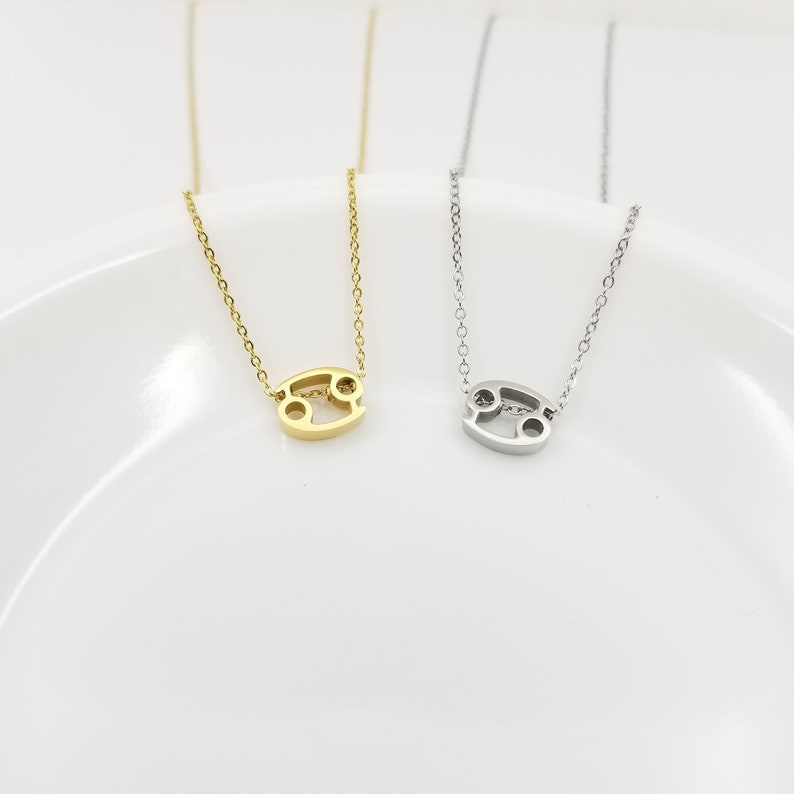 May include: Two delicate gold and silver necklaces with a Cancer zodiac sign pendant. The necklaces are displayed on a white plate.