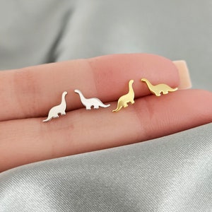 May include: Three pairs of small dinosaur earrings. One pair is gold, one pair is silver, and one pair is a mix of gold and silver.