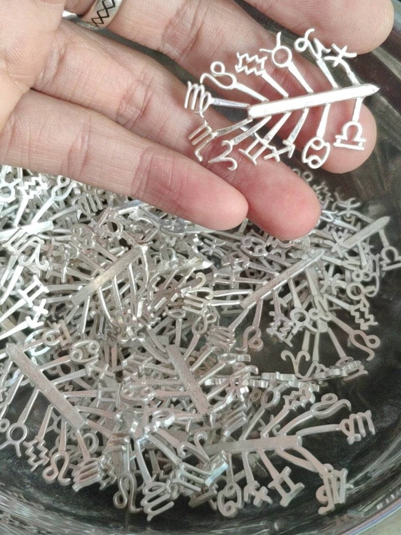 May include: A close-up of a pile of silver zodiac sign charms. The charms are in the shape of the twelve astrological signs. The charms are arranged in a random order.
