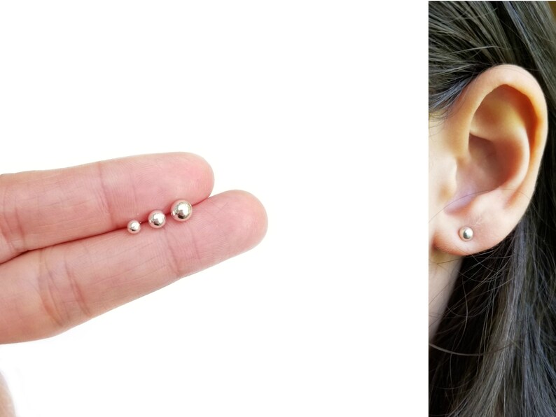 May include: Three silver ball stud earrings of different sizes. The earrings are shown on a hand and in a person's ear.