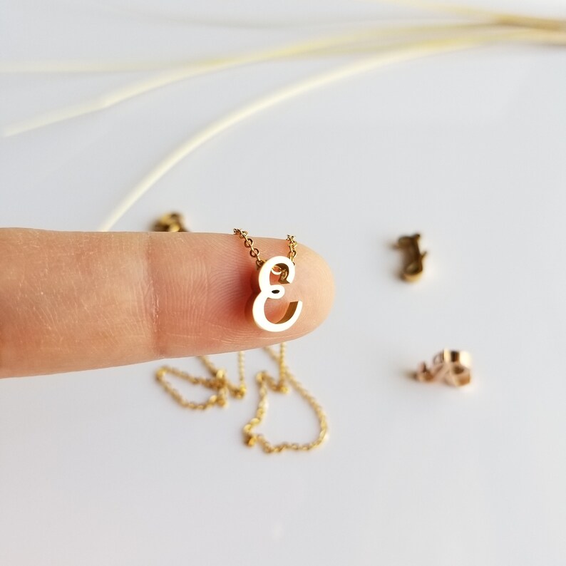 May include: A gold chain necklace with a small, cursive letter 'E' pendant. The pendant is held by a finger.