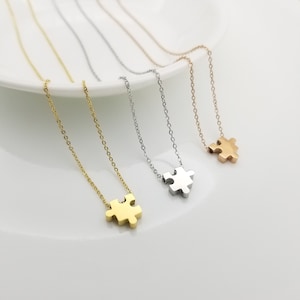 May include: Four silver necklaces with puzzle piece pendants. The necklaces are on a white surface. The puzzle pieces are in gold, silver, and rose gold.