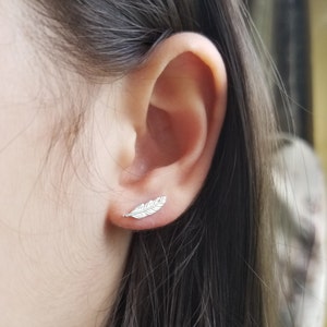May include: A close-up of a person's ear with a small silver feather earring. The earring is a simple design with a delicate feather shape.