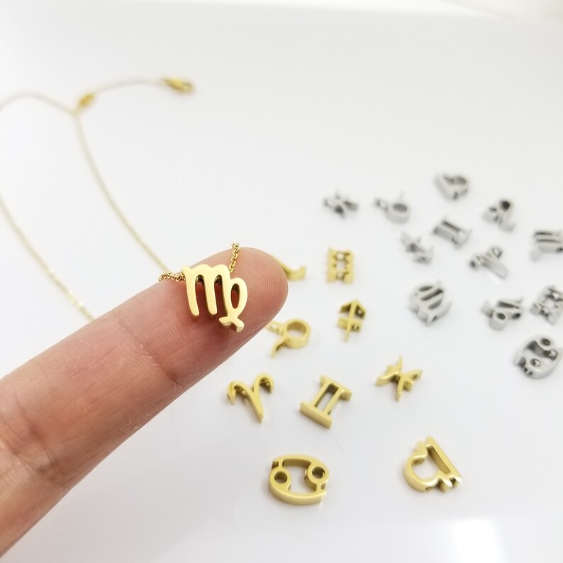 May include: A gold necklace with a small Virgo zodiac sign pendant. The pendant is on a finger, and there are other zodiac signs scattered around it on a white surface.