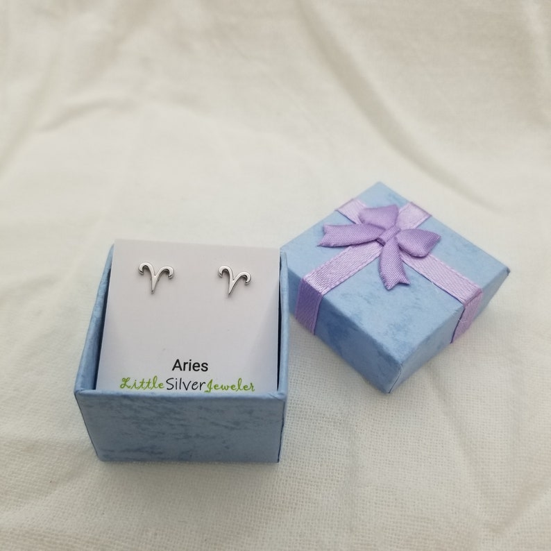 May include: Silver Aries zodiac sign earrings in a light blue gift box with a purple ribbon. The box has the text "Aries LittleSilverjeweler" on the front.