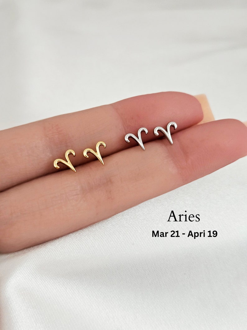 May include: A pair of gold and silver earrings featuring the zodiac symbol for Aries. The earrings are shown on a person's finger. The text "Aries Mar 21 - Apr 19" is below the earrings.