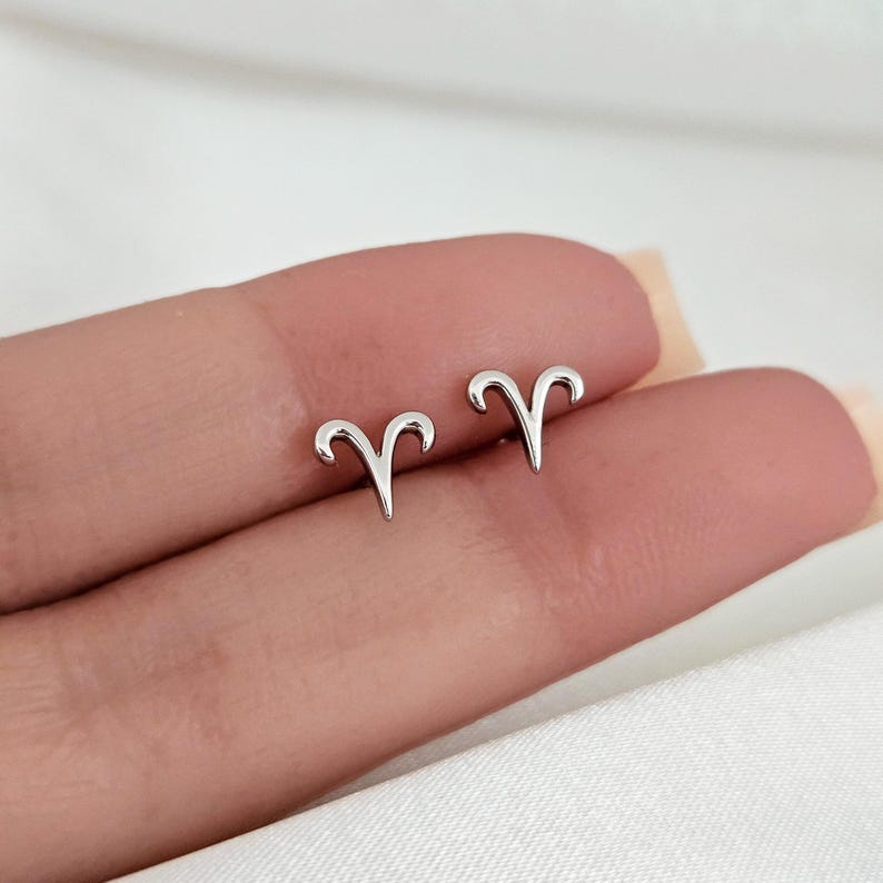 May include: A pair of silver Aries zodiac sign stud earrings. The earrings are small and delicate, and they are perfect for everyday wear.