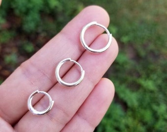 May include: Three small, silver hoop earrings are shown against a blurred green backdrop. The earrings are round with a simple, polished design. The earrings are held in a person's hand.