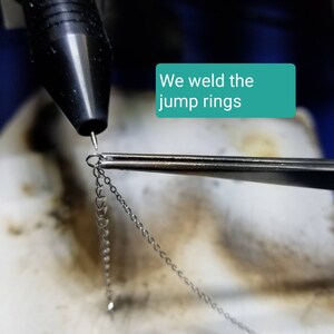 May include: A close-up of a silver jump ring being welded with a torch. The jump ring is held in place with tweezers. The text "We weld the jump rings" is visible in the image.