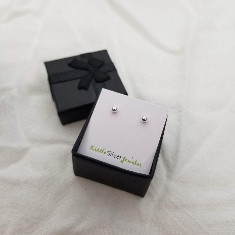 May include: A pair of small, silver stud earrings in a black gift box with a white insert. The insert has the text "Little Silver Jeweler" printed on it.