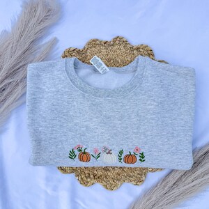 May include: A light gray sweatshirt with an embroidered design of pumpkins and flowers. The design includes two orange pumpkins, one white pumpkin, and pink flowers. The sweatshirt is made of a soft material and is perfect for fall.