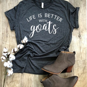 May include: A dark gray t-shirt with the text "Life is better with goats" printed in white. The shirt is laying on a wooden surface with a sprig of cotton and a pair of brown ankle boots.
