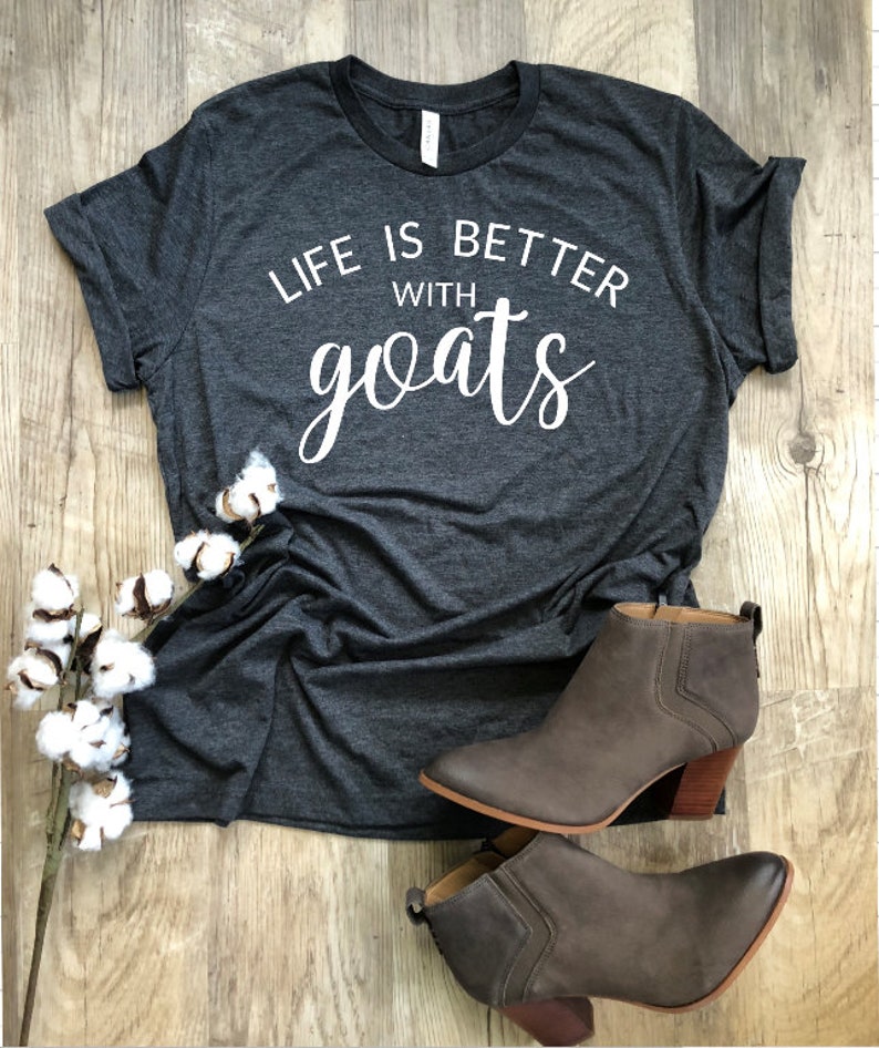 May include: A dark gray t-shirt with the text "Life is better with goats" printed in white. The shirt is laying on a wooden surface with a sprig of cotton and a pair of brown ankle boots.