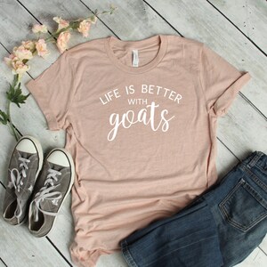 May include: A light pink t-shirt with the text "Life is better with goats" in white lettering. The shirt is laid out on a wooden surface with a pair of gray sneakers and blue jeans.