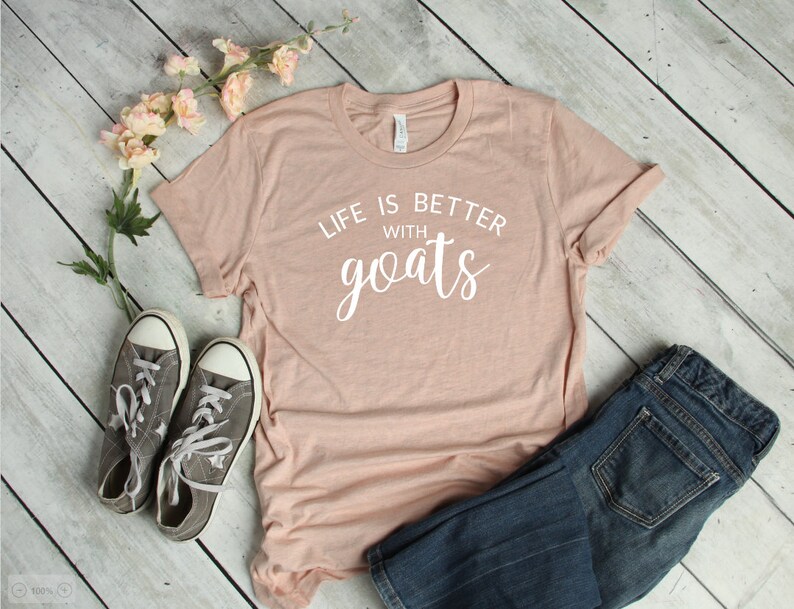 May include: A light pink t-shirt with the text "Life is better with goats" in white lettering. The shirt is laid out on a wooden surface with a pair of gray sneakers and blue jeans.