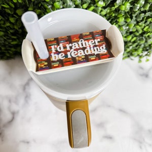 Peut inclure: Une tasse &agrave; emporter blanche avec une poign&eacute;e marron et un couvercle gris. Le couvercle a un autocollant qui dit "J'aimerais mieux lire" avec un fond de livres color&eacute;s.