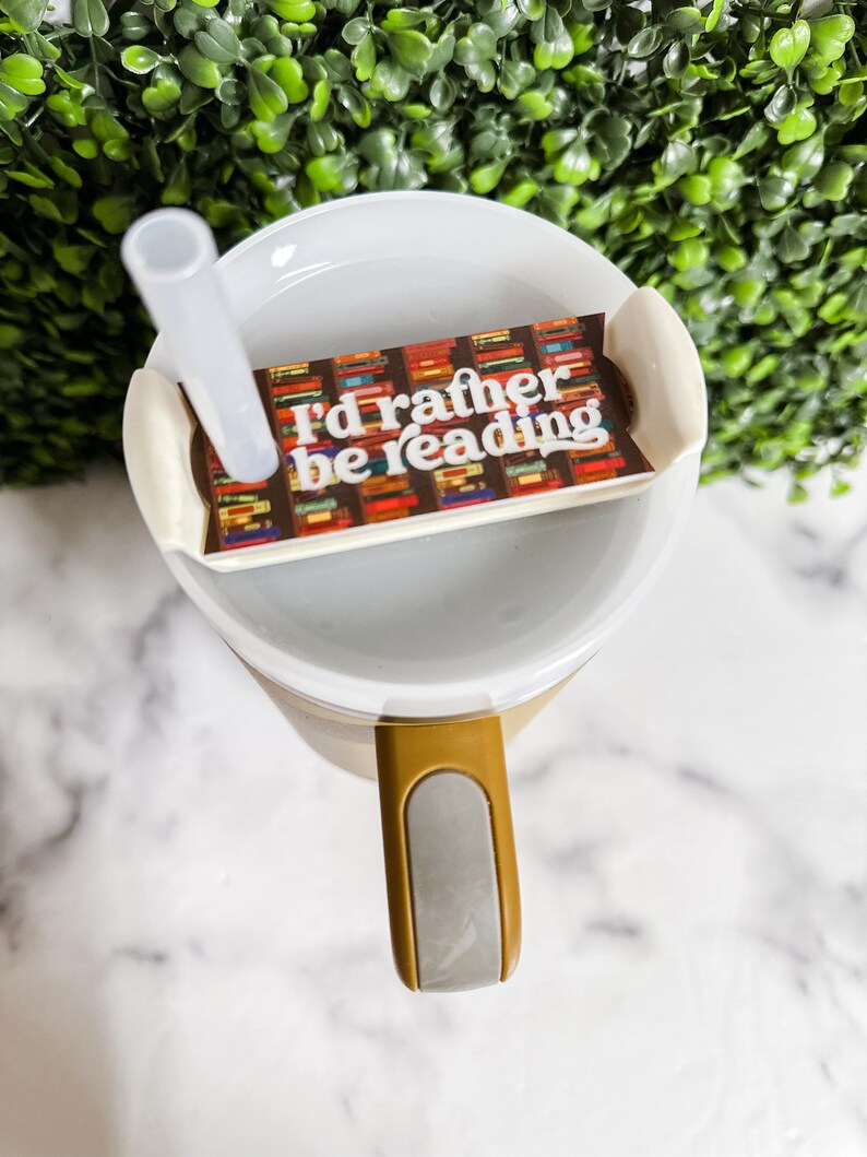 Peut inclure: Une tasse &agrave; emporter blanche avec une poign&eacute;e marron et un couvercle gris. Le couvercle a un autocollant qui dit "J'aimerais mieux lire" avec un fond de livres color&eacute;s.