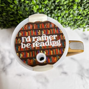 Peut inclure: Mug de voyage blanc avec un couvercle et une poign&eacute;e dor&eacute;s. Le mug est d&eacute;cor&eacute; d'un graphique repr&eacute;sentant une biblioth&egrave;que remplie de livres color&eacute;s et le texte "I'd rather be reading".