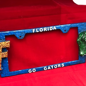 May include: Blue and white license plate frame with the words "Florida" and "Go Gators". A gold glitter "F" is on the left side and a green alligator head is on the right side.