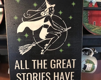 May include: Black wooden sign with a white illustration of a witch on a broomstick, surrounded by green stars. The sign reads "ALL THE GREAT STORIES HAVE Witches IN THEM" in white and green lettering. Decorative Halloween theme.