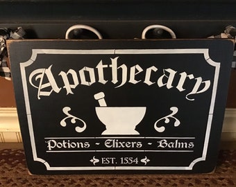 May include: Black rectangular sign with white lettering and a white border. The sign reads "Apothecary" above an image of a mortar and pestle. Below the image, it reads "Potions - Elixers - Balms" and "EST. 1554". Two white handles are at the top.