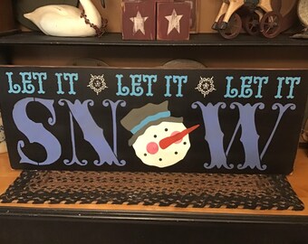 May include: A rectangular wooden sign with the words "LET IT SNOW" in blue and white lettering. The "SNOW" features a snowman graphic with a red carrot nose and a gray hat. The background is black.