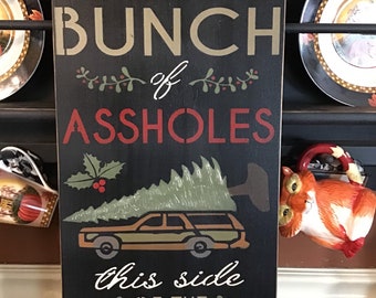 May include: A rectangular black wooden sign with white, tan, and red lettering. The sign reads "HOME OF THE JOLLIEST BUNCH of ASSHOLES" and "this side OF THE NUTHOUSE". A station wagon with a Christmas tree is also depicted.