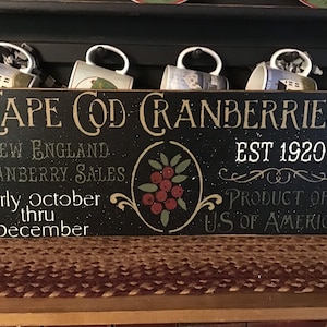 May include: A black wooden sign with the words "Cape Cod Cranberries" and "EST 1920" in white lettering. The sign also includes the text "New England Cranberry Sales" and "Product of US of America". Four mugs are displayed above the sign.