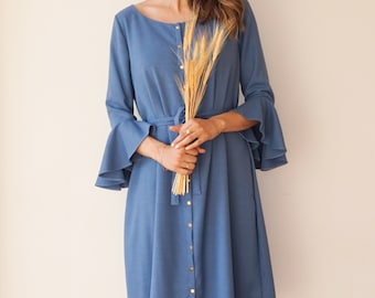 May include: A blue dress with a button-down front, long sleeves with flared cuffs, and a matching belt. The dress is knee-length and paired with dark blue heels. The model is holding a bundle of wheat.